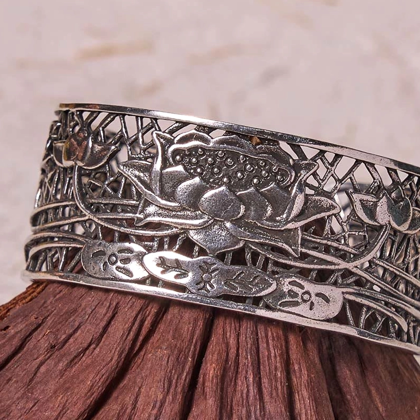 Low angle side view of silver plated hollow lotus bracelet with large central lotus carving and decorative borders on wooden texture base