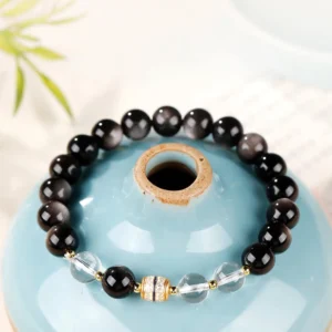 Silver Sheen Obsidian and Dzi Bead Buddhist protection bracelet with black beads and clear quartz spacers, displayed on blue ceramic jar