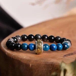Silver Sheen Obsidian Blue Tiger Eye Buddhist protection bracelet with engraved Om Mani Padme Hum copper charm, overhead top-down view on wooden surface