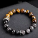 Silver Sheen Obsidian and Tiger's Eye Buddhist bracelet with engraved Om Mani Padme Hum charm on dark display board, showing intricate metal spacer beads and two-tone stone arrangement