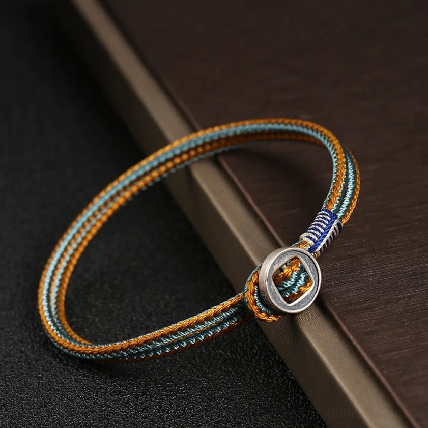 Single-strand woven Buddhist protection bracelet with brown cyan-blue cord and round metal buckle, side view on wooden board