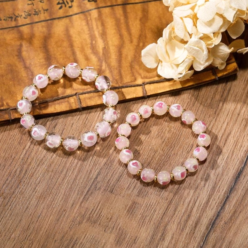 Pair of plain pink translucent luminous glass bead Buddhist bracelets with gold spacer beads arranged flat lay on wooden table with dried flowers
