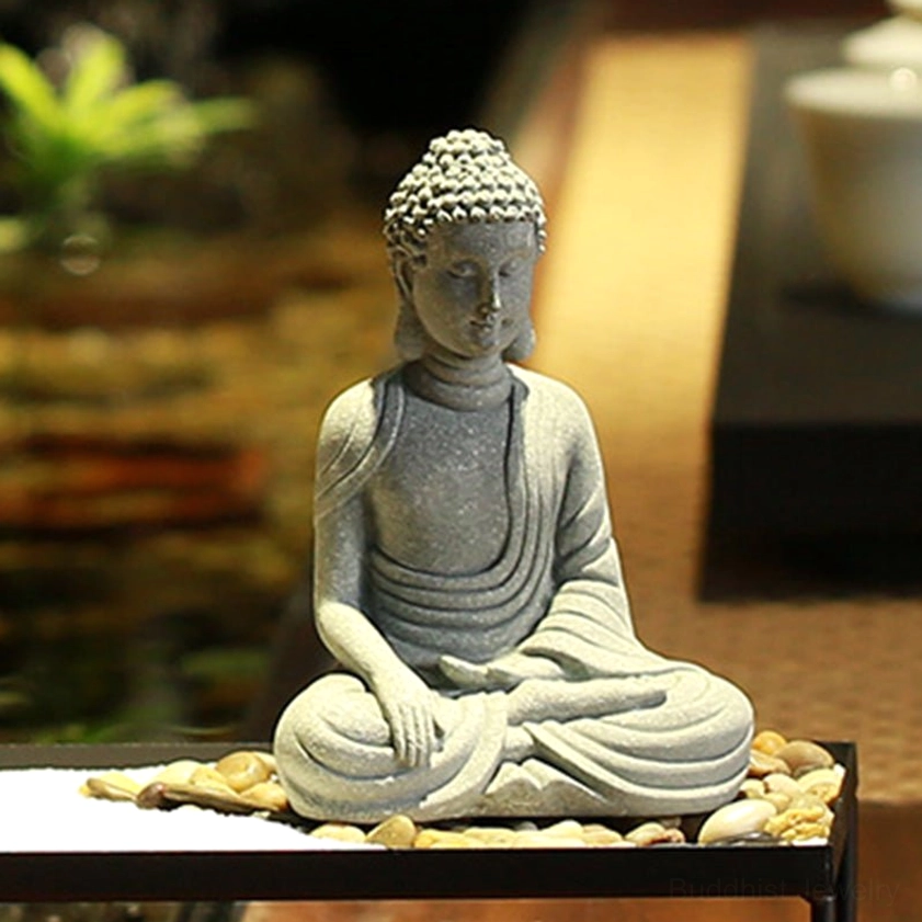Grey matte sandstone Buddha meditation statue displayed on zen table with indoor greenery and natural lighting, front three-quarter perspective