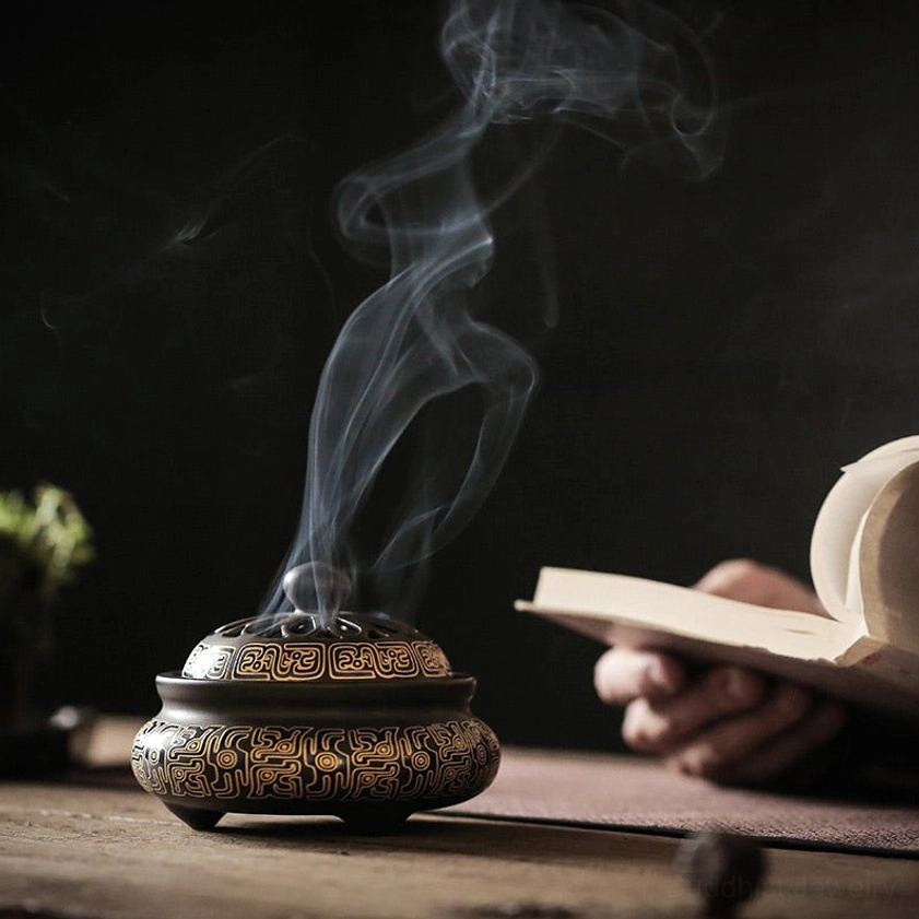 Small dark brown ceramic incense burner with floral patterns, hollow lid, rising incense smoke, lifestyle scene with book