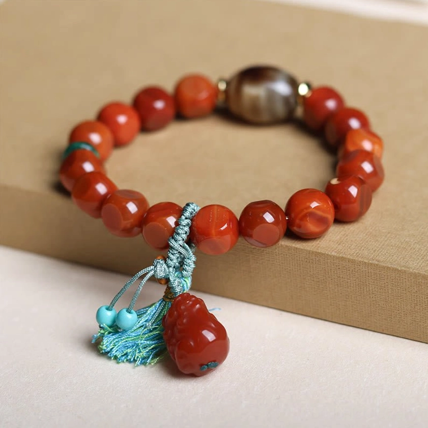 South Red Agate Pixiu wealth bracelet with faceted beads, teal tassel charm, and brown central bead on cardboard base