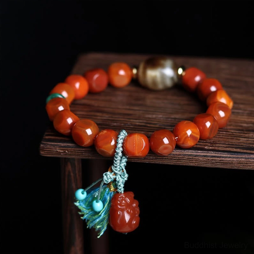 South Red Agate Pixiu bracelet with teal tassel charm, faceted beads on wooden display stand showing three-quarter view