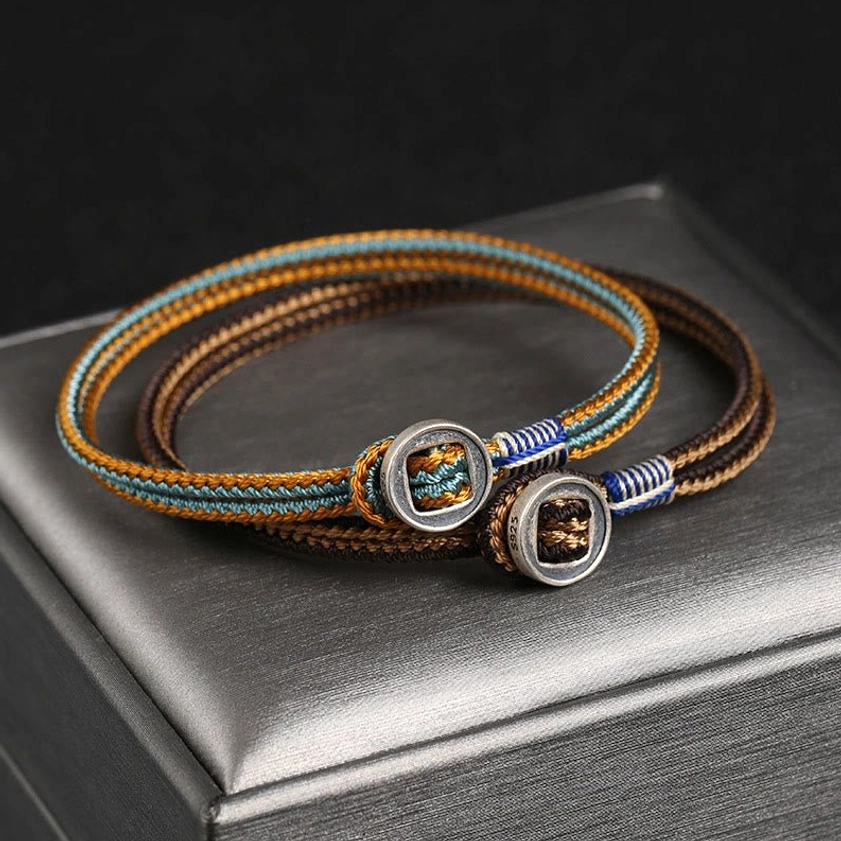 Handmade dual-strand silver string bracelet with brown blue woven cord and metal buttons, Buddhist protection design displayed on jewelry box
