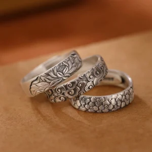 Three handcrafted 999 sterling silver Buddhist rings with engraved lotus, plum blossom, and auspicious cloud designs displayed on brown leather, top-down flat lay view