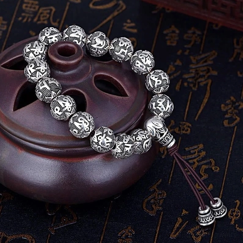 Dark brown cord mala bracelet with silver carved mantra beads and double tassel clasp on dark teapot background