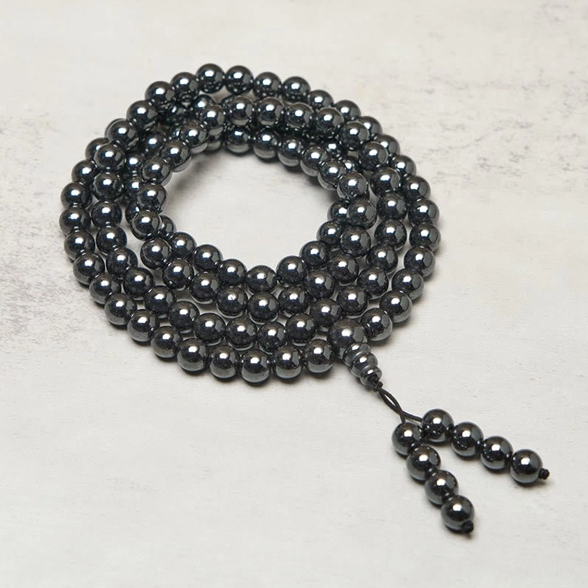 108 bead Terahertz mala prayer beads coiled on light neutral surface, dark metallic polished beads with tassel pendant