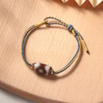 Three-eye Dzi bead bracelet flat lay top view with blue and gold braided cord on wooden surface