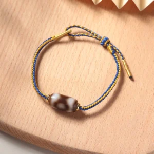 Three-eye Dzi bead bracelet flat lay top view with blue and gold braided cord on wooden surface