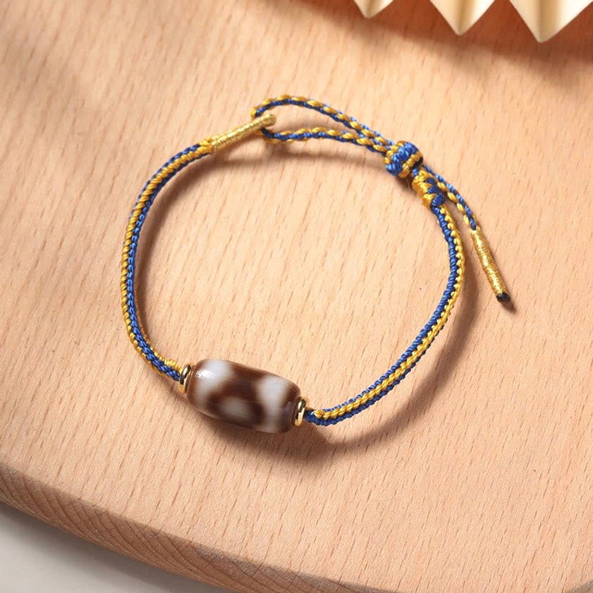 Three-eye Dzi bead bracelet flat lay top view with blue and gold braided cord on wooden surface
