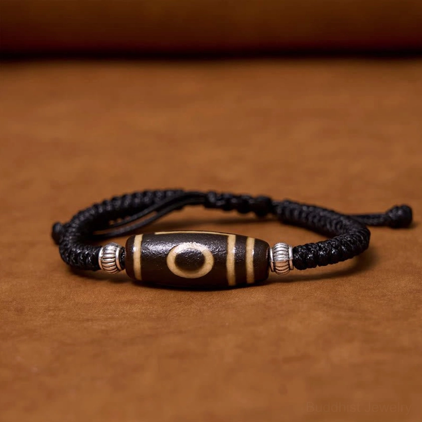 Three-eye dzi bead bracelet flat lay on brown wooden background showing three-eye pattern design with black cord