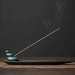 Three Stones ceramic incense holder with lit incense stick on dark wooden table, showing stacked stone design and matte teal glaze