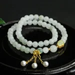 Tianshan jade lotus double circle bracelet with pearl pendants on black ceramic tray, top view showing pale green jade beads and gold clasp