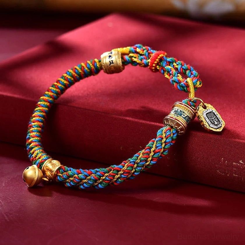 Buddhist amulet bracelet front close-up with multicolor woven rope, gold metal parts, bell and protective tag pendants on red silk