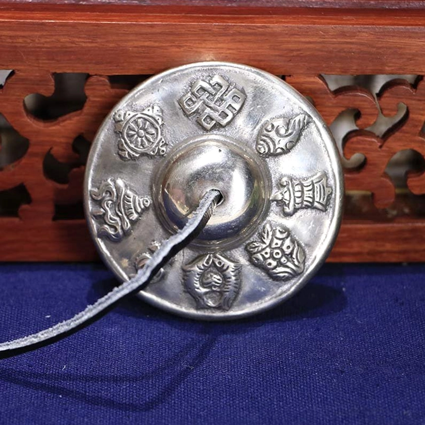 Single Buddhist tingsha bell with eight auspicious symbols embossed, polished white copper, dark blue background, flat view
