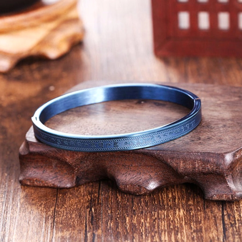 Burnt blue titanium steel Buddhist blessing bangle with smooth matte surface and engraved band edges on carved wooden display