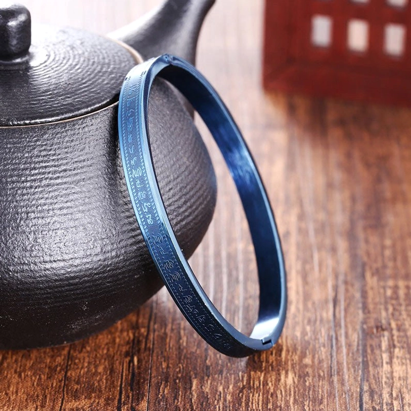 Matte blue titanium steel blessing bracelet shown from oblique side view next to traditional black ceramic teapot on wooden table