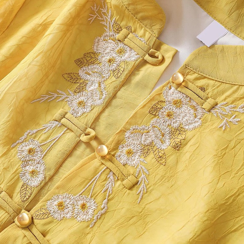 Side detail view of yellow tunic front placket showing traditional frog button fasteners with golden beads and intricate white floral embroidery on wrinkled fabric