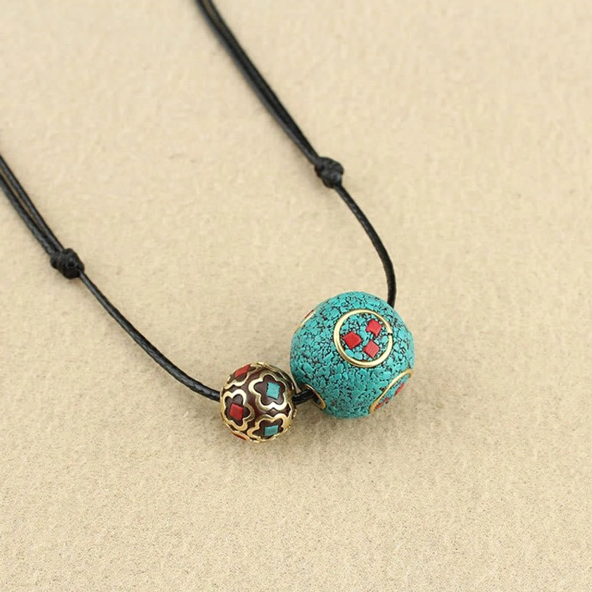 Handcrafted turquoise and metal bead Buddhist necklace with black cord, oblique side view on beige tabletop