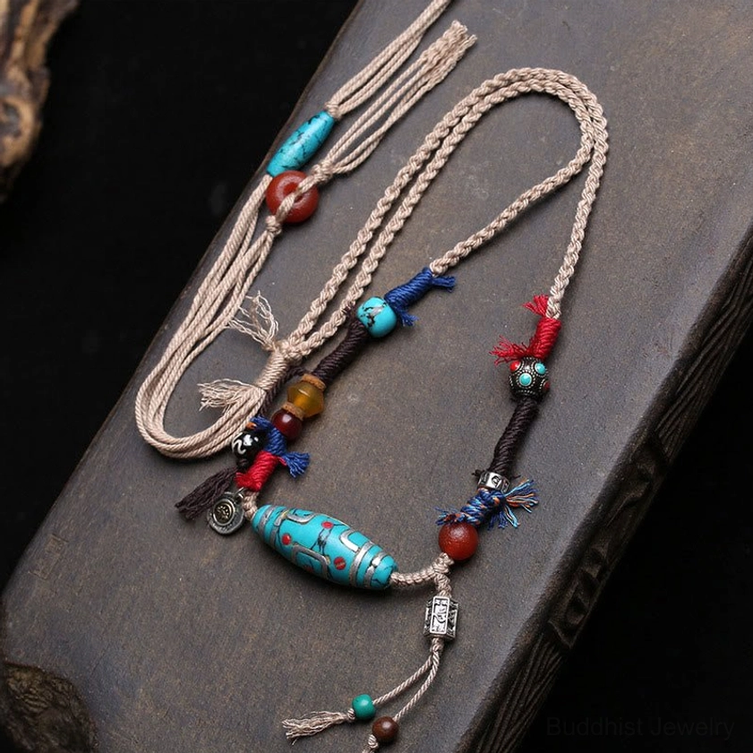 Turquoise dzi bead copper necklace pendant with braided cotton cord on dark wooden surface, showing complete Buddhist protection amulet design