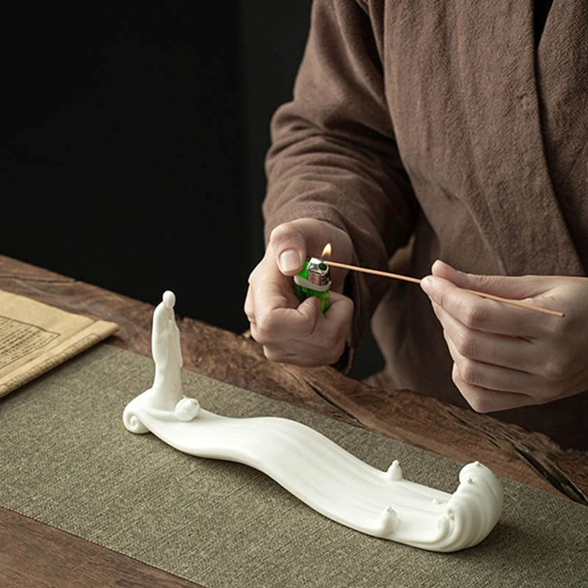 Hand placing incense stick into white ceramic Buddhist burner on tea table with linen mat