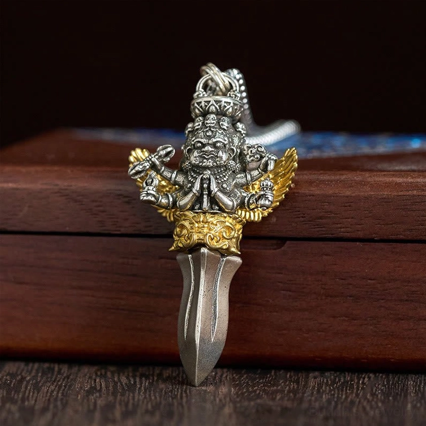 Six-armed dharma protector vajra pendant close-up front view on wooden box background, showing deity figure in prayer gesture with intricate metal blade details