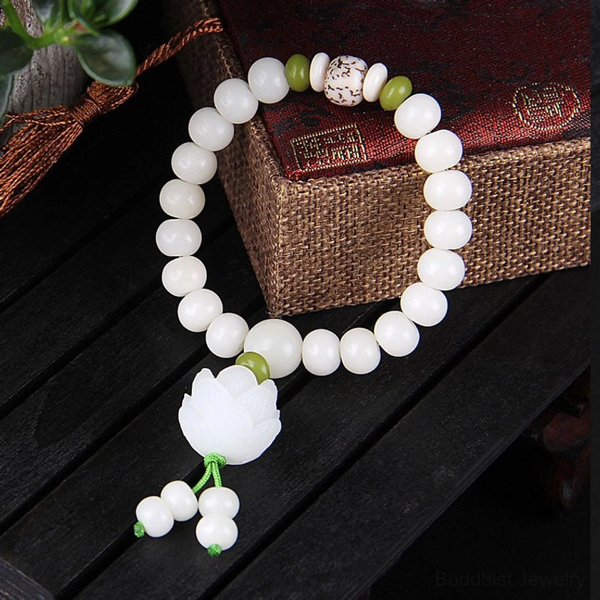 Bodhi seed meditation bracelet side overhead view with smooth white beads, green spacers, and white lotus tassel charm next to linen gift box on dark wood table