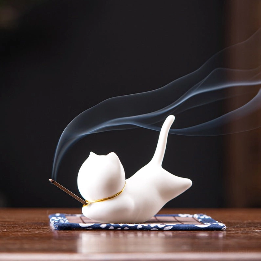 White cat ceramic incense holder with double incense sticks creating smoke, gold accent collar detail, front view on polished wooden table