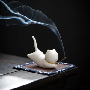 White ceramic cat incense burner with gold collar, burning incense on dark wood surface, showing active smoke trails from meditation practice