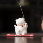 White ceramic lucky cat incense holder with red collar and gold bell, front view on dark wooden desk
