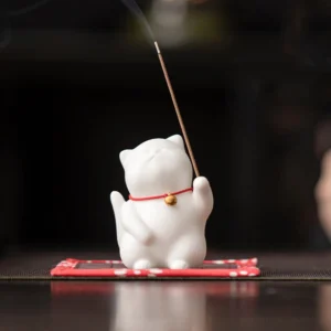 White ceramic lucky cat incense holder with red collar and gold bell, front view on dark wooden desk