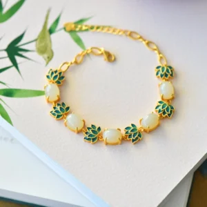 White jade and cyan jade lotus Buddhist bracelet with gold-toned chain, displayed overhead on a book with bamboo leaf decoration