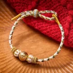 White braided rope Om Mani Padme Hum wisdom bracelet with gold-toned mantra bead and decorative knot, top-down flat lay view