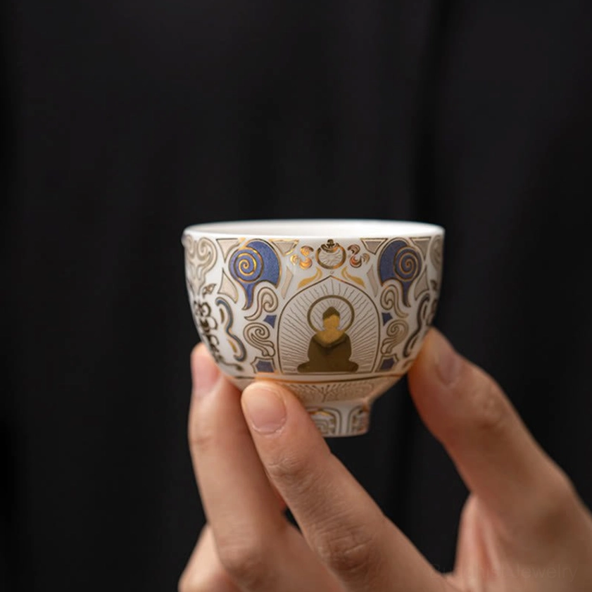 Authentic handmade ceramic kung fu tea cup with Buddhist motif, held by two hands against plain dark background, showing detailed Buddha illustration