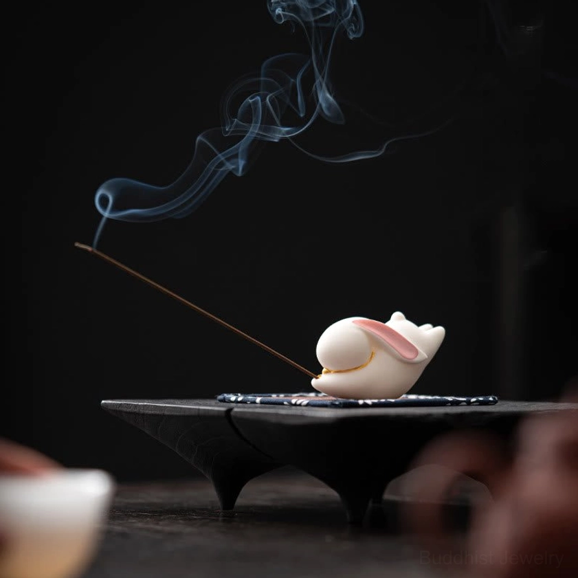White ceramic rabbit incense holder with burning incense and smoke on black pedestal base