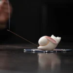 White ceramic rabbit incense burner with pink inner ears and gold trim on blue floral bamboo mat with burning incense stick