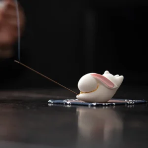 White ceramic rabbit incense burner with pink inner ears and gold trim on blue floral bamboo mat with burning incense stick