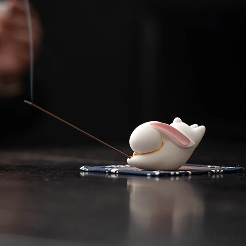 White ceramic rabbit incense burner with pink inner ears and gold trim on blue floral bamboo mat with burning incense stick