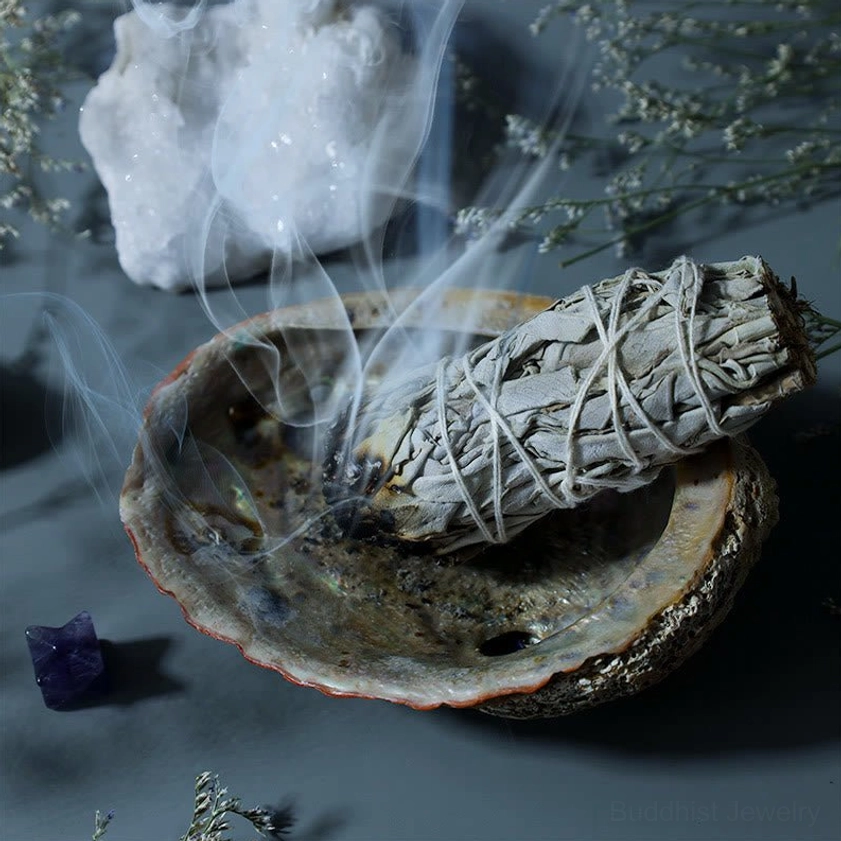 Lit white sage smudge stick resting in abalone shell holder with rising smoke and dried flowers for meditation
