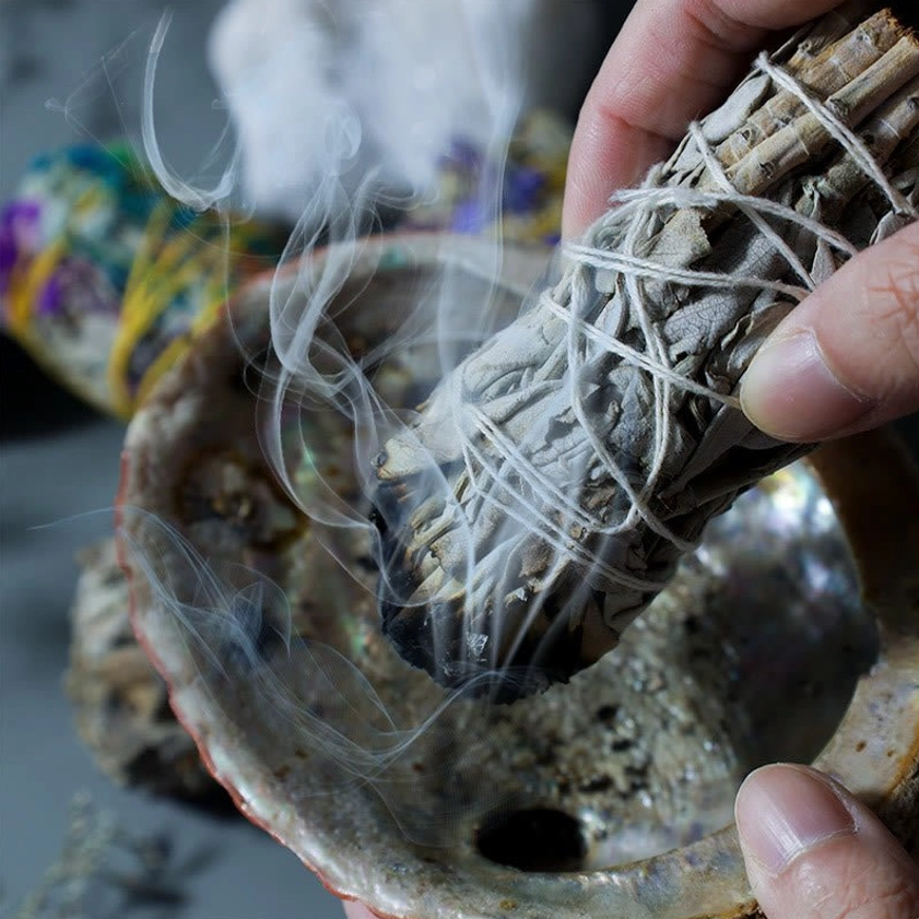 White sage smudge stick held over abalone shell incense burner with active smoke for space cleansing