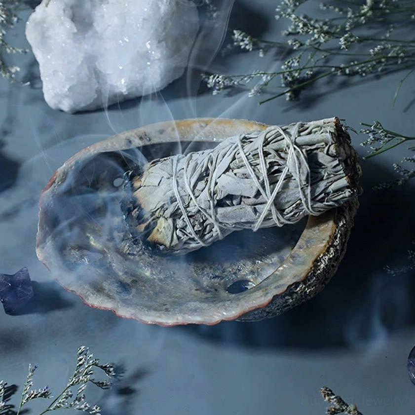 Smoldering white sage smudge stick in abalone shell tray with clear quartz and dried flowers for spiritual practice