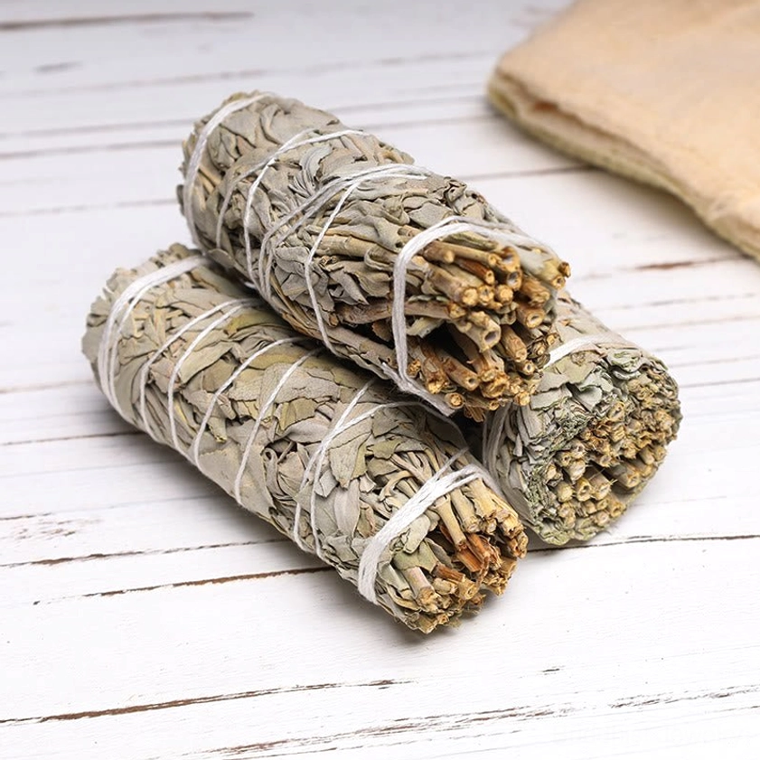 Three white sage smudge sticks arranged on white wooden surface with burlap cloth for spiritual cleansing rituals