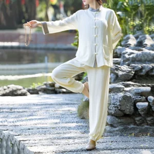 Women's beige cotton linen tai chi meditation suit with frog buttons, side view standing on stone steps by water