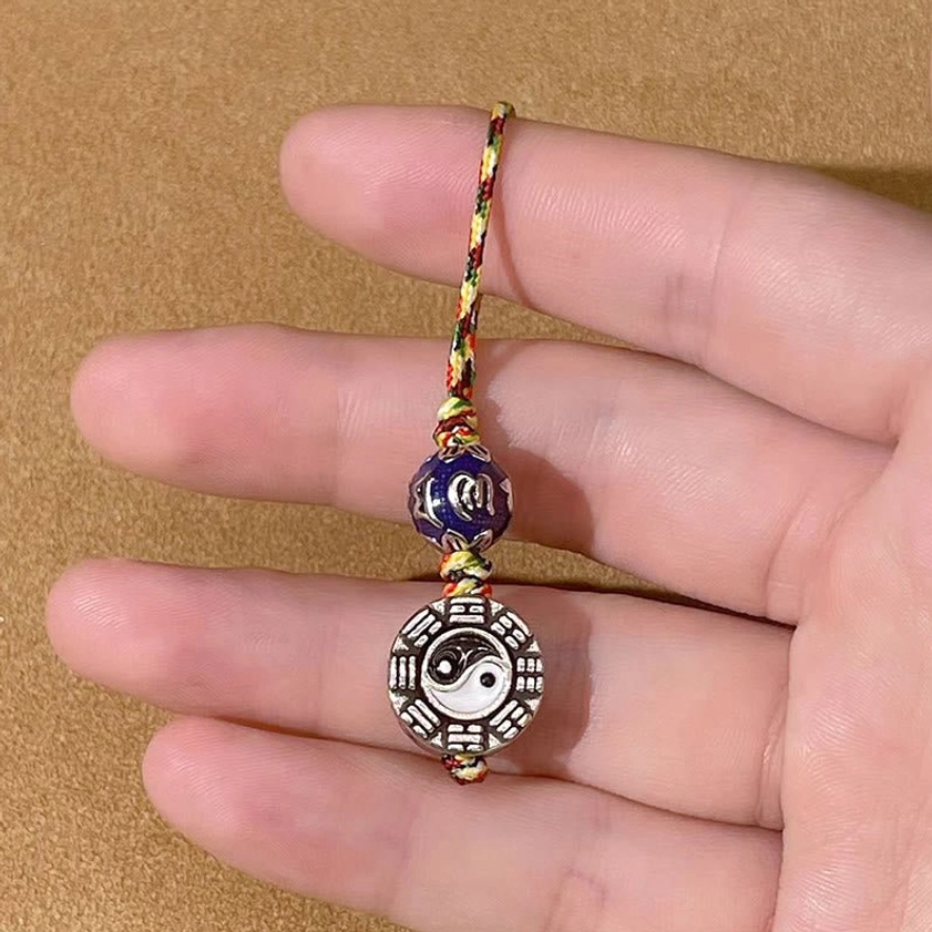 Hand-held view of sacred Buddhist keychain showing intricate Bagua Yin Yang pendant with dark blue enamel bead and braided lanyard