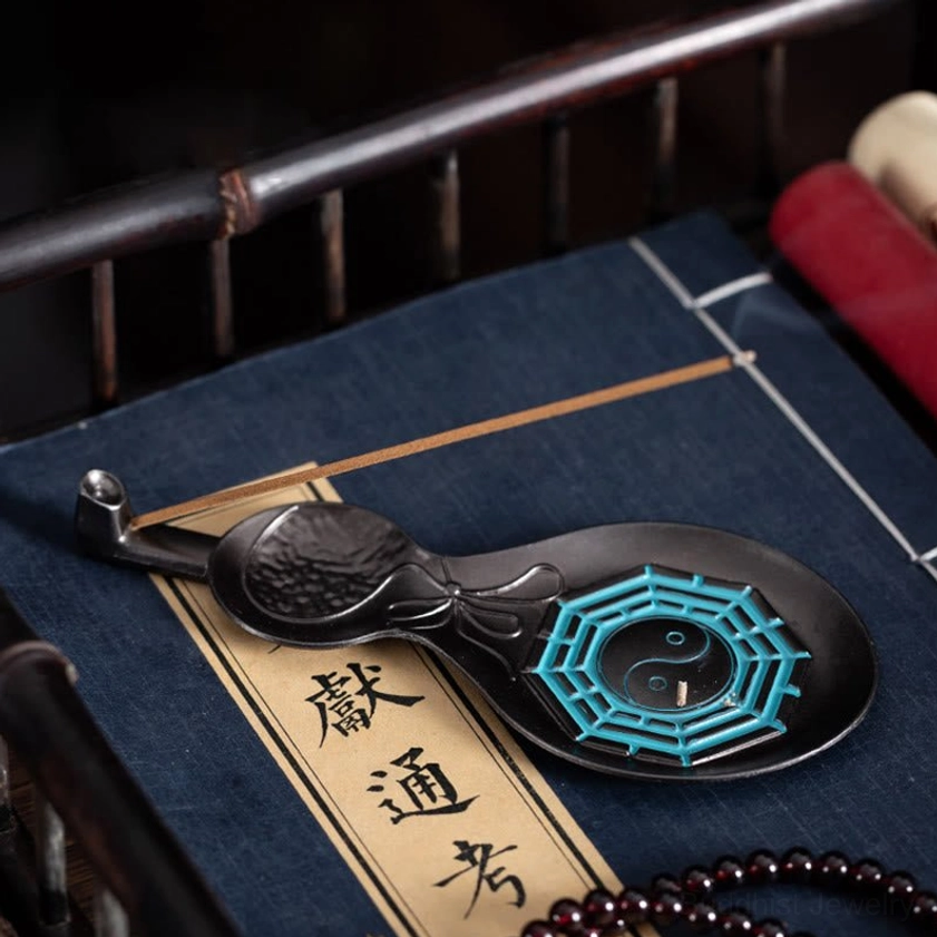 Black copper Yin Yang Bagua incense holder displaying cyan patterns surrounded by Buddhist prayer beads and ancient spiritual texts