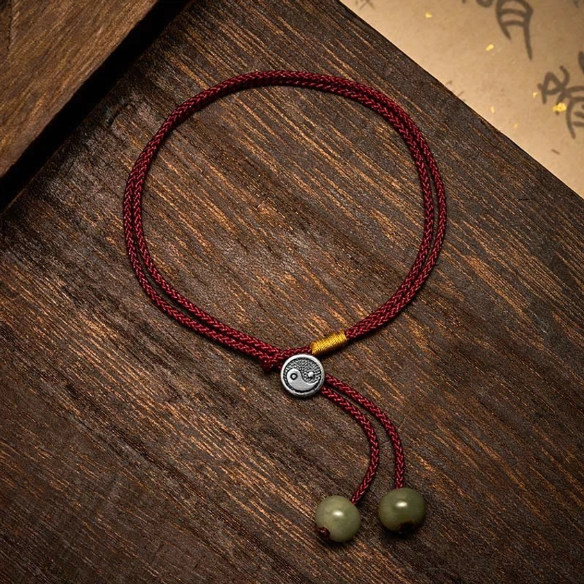 Top-down flat lay view of Yin Yang protection bracelet showing braided red cord, silver charm detail, and dual green bead tassel closure