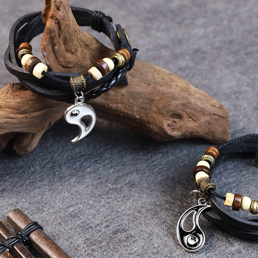 Yin yang couple bracelets with layered leather, wooden accent beads, and split metal charms displayed on driftwood desktop